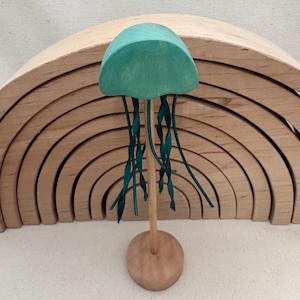 May include: A wooden jellyfish toy with a teal cap and dark teal tendrils. The jellyfish is mounted on a wooden stand. A wooden rainbow arch is in the background. The toy is designed for children.