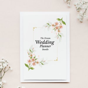 May include: A white wedding planner bundle with the text "The Dream Wedding Planner Bundle" in black. The planner is decorated with watercolor floral designs in pink, green, and gold. White flowers are in the background.
