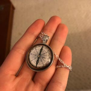 May include: Silver compass pendant necklace with a detailed compass rose design. The compass face is white with black directional markings and a silver chain. The pendant is round and has a silver bezel.