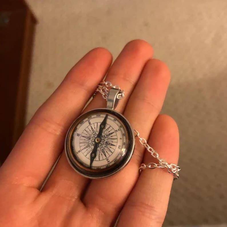 May include: Silver compass pendant necklace with a detailed compass rose design. The compass face is white with black directional markings and a silver chain. The pendant is round and has a silver bezel.