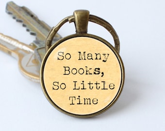 Puede incluir: Un llavero de bronce con un dije redondo de vidrio. El dije tiene un fondo beige con texto negro que dice "So Many Books, So Little Time".