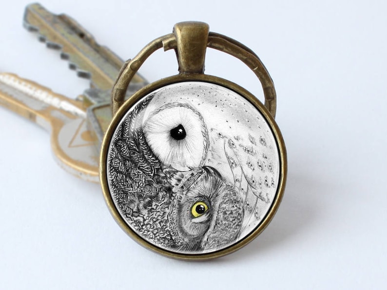 May include: A round glass keychain with a bronze metal frame features a black and white yin yang design with two owls facing each other. The owl on the left has a yellow eye and the owl on the right has a black eye.