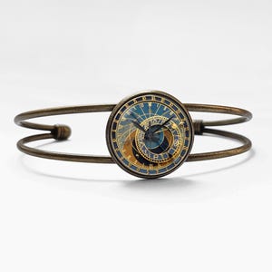 Puede incluir: Pulsera de puño en tono bronce con una esfera redonda de reloj con un diseño de reloj astronómico azul y dorado. La pulsera tiene un diseño sencillo y abierto con una bisagra. Un accesorio único.