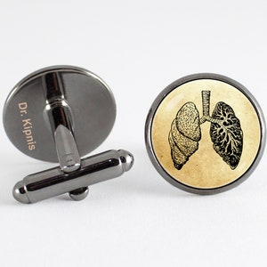 May include: A pair of cufflinks with a black metal base and a round glass top. The glass top features a vintage illustration of a human lung. One cufflink has the text "Dr. Kipnis" engraved on the metal base.