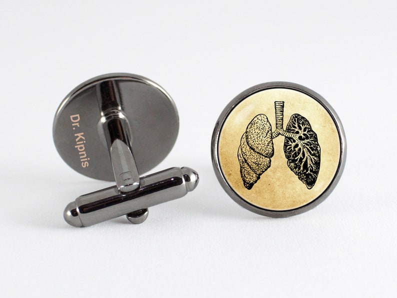 May include: A pair of cufflinks with a black metal base and a round glass top. The glass top features a vintage illustration of a human lung. One cufflink has the text "Dr. Kipnis" engraved on the metal base.