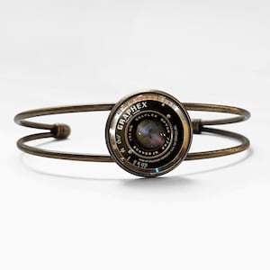 Peut inclure: Un bracelet manchette de couleur bronze avec un objectif d'appareil photo Graphex Optar vintage en son centre. L'objectif est noir et argenté avec des marquages détaillés. Le bracelet a un design simple et ouvert.