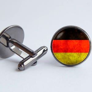May include: A pair of cufflinks with a black metal base and a round glass top featuring the German flag. The flag is red, yellow, and black with a distressed look.