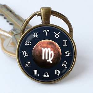 Op de afbeelding: Een bronzen sleutelhanger met een ronde glazen hanger met een donkerblauwe achtergrond met witte dierenriemsymbolen en een rood-oranje planeet met het dierenriemteken Maagd in wit.