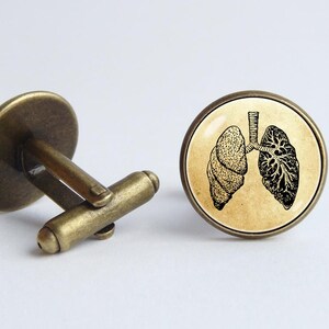 May include: A pair of cufflinks with a vintage illustration of human lungs on a beige background. The cufflinks are made of brass and have a round shape.