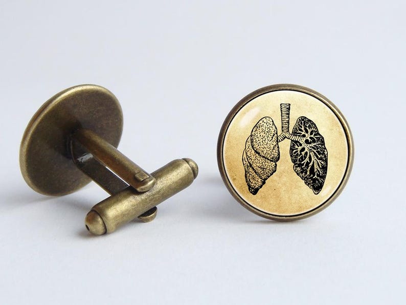 May include: A pair of cufflinks with a vintage illustration of human lungs on a beige background. The cufflinks are made of brass and have a round shape.