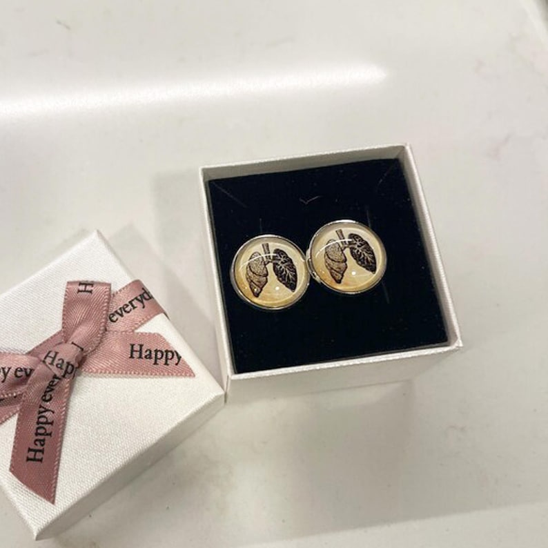 May include: A pair of cufflinks in a white box with a pink ribbon. The cufflinks are round and have a silver finish. The design on the cufflinks is a detailed image of a pair of lungs.