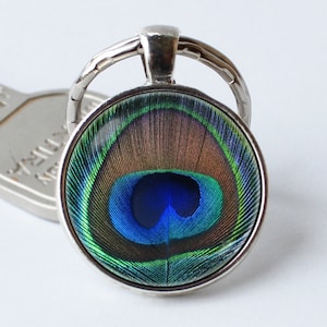 以下が含まれることがあります： 青、緑、茶色の孔雀の羽根が描かれた丸いガラス製のチャームが付いたシルバーのキーホルダーです。