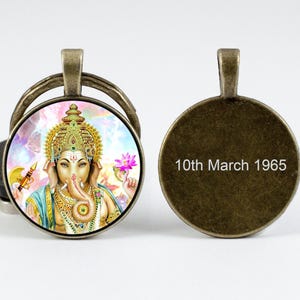 May include: A bronze key chain with a round glass charm featuring a colorful image of the Hindu deity Ganesha. The back of the charm is blank and engraved with the text "10th March 1965".