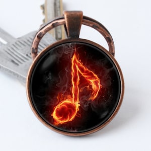 Puede incluir: Un llavero de color cobre con un dije redondo de vidrio que presenta una nota musical roja fuego sobre un fondo negro.