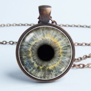 Puede incluir: Un collar de cadena de bronce con un colgante redondo que presenta una imagen de primer plano de un ojo humano con un iris gris y una pupila negra.