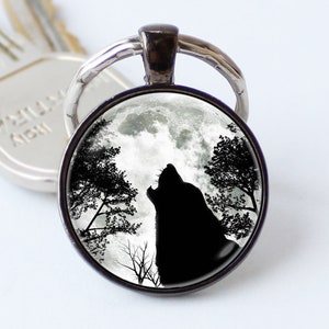 Puede incluir: Un llavero plateado con un dije redondo de vidrio que presenta una silueta negra de un lobo aullando a la luna. La luna está llena y blanca con nubes en el fondo.