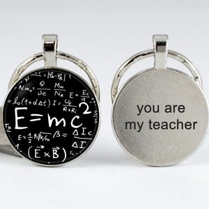 May include: A silver keychain with two round charms. One charm features the equation E=mc² written on a black background. The other charm has the text "you are my teacher" on a silver background.