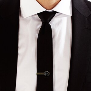 Puede incluir: Una corbata negra con un clip de corbata plateado que dice "0.6" en una camisa blanca y una chaqueta de traje negra.