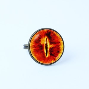May include: A ring with a red and orange eye design. The ring is made of metal and has a round shape.