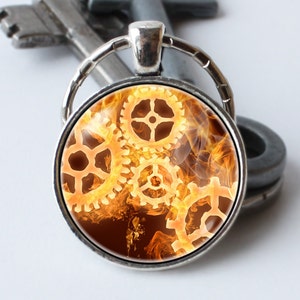 May include: A silver key chain with a round glass charm featuring a design of interlocking gears set against a background of orange and yellow flames.