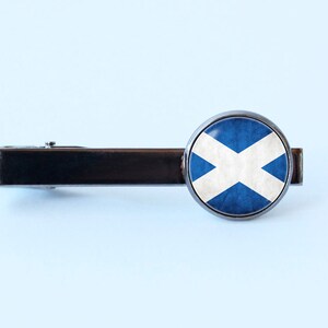 Puede incluir: Un pasador de corbata plateado con un dise&ntilde;o redondo de bandera escocesa azul y blanca.