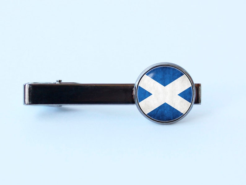 Puede incluir: Un pasador de corbata plateado con un dise&ntilde;o redondo de bandera escocesa azul y blanca.