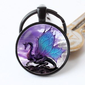 Peut inclure: Un porte-clés en métal noir avec un charme rond en verre représentant un dragon violet et bleu avec des ailes déployées sur un fond de ciel violet nuageux.