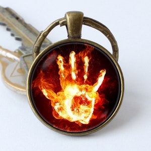 May include: A bronze key chain with a round glass charm featuring a white hand engulfed in orange and red flames.