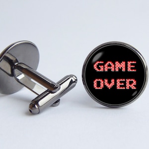 Può includere: Gemelli color canna di fucile con una faccia nera con la scritta "GAME OVER" in rosso pixelato. I gemelli hanno un design classico con una faccia arrotondata.