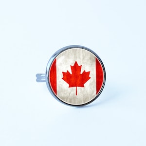 Puede incluir: Un anillo de plata con un diseño redondo de bandera canadiense blanca y roja. La bandera tiene una hoja de arce roja sobre un fondo blanco con una franja roja a cada lado.