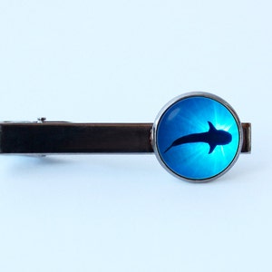 May include: A silver tie bar with a round blue glass cabochon featuring a silhouette of a whale shark swimming in blue water.