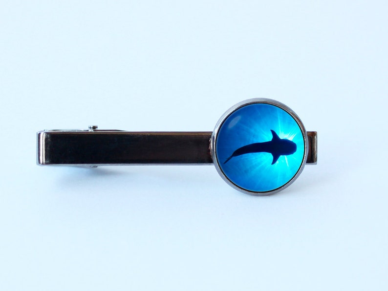 May include: A silver tie bar with a round blue glass cabochon featuring a silhouette of a whale shark swimming in blue water.
