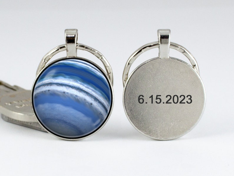 May include: A silver key chain with a round blue and white agate stone on one side and the date 6.15.2023 engraved on the other side.