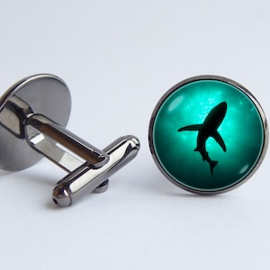 Op de afbeelding: Een paar manchetknopen met een zwarte metalen basis en een ronde glazen bovenkant. De glazen bovenkant heeft een silhouet van een haai die zwemt in een turquoise blauwe achtergrond.