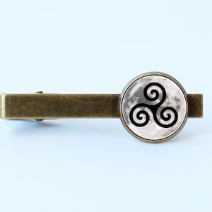 May include: A bronze tie bar with a round glass cabochon featuring a black and white image of a triskelion symbol on a moon background.