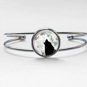 May include: Silver-tone cuff bracelet featuring a round, black and white image of a howling wolf against a full moon. The bracelet has a double-band design and is made of a shiny, metallic material. The overall theme is nature and wildlife.