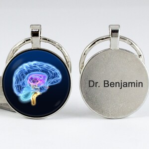 May include: A silver keychain with a round glass charm featuring a blue and pink anatomical brain illustration. The other side of the keychain has the text "Dr. Benjamin" engraved on it.