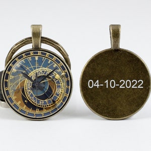 May include: A round, antique gold-toned pendant with a detailed astronomical clock face on one side and the date "04-10-2022" engraved on the other side.