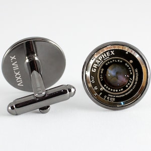 Puede incluir: Un par de gemelos de plata con fondo negro. Un gemelo presenta la inscripción en números romanos "X.VII.XXIV". El otro gemelo presenta una lente de cámara vintage con el texto "GRAFLEX TRADE MARK GRAFLEX OPTAR 101 mm 1:4.5 5.6 8 11 16 2232 MADE BY WOLLENSAK ROCHESTER USA FOR GRAFLEX INC. 20 25 30 40 50 100 200 400".