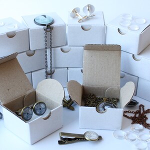 May include: Assortment of white cardboard boxes containing various jewelry items, including necklaces, cufflinks, and tie clips. The boxes are arranged in a pyramid shape on a white surface. Some of the jewelry items are visible in the boxes, including a necklace with a glass pendant, cufflinks with a glass design, and a tie clip with a glass design.
