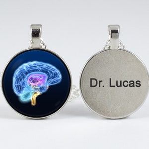 Peut inclure: Un collier en argent avec un pendentif rond représentant une illustration de cerveau bleu et violet d'un côté et le texte "Dr. Lucas" de l'autre.