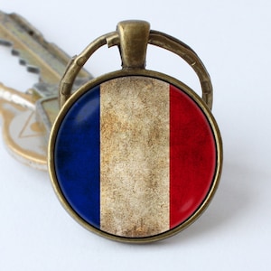 Puede incluir: Un llavero redondo de color bronce con un inserto de vidrio que presenta una bandera francesa desgastada. La bandera es azul, blanca y roja.