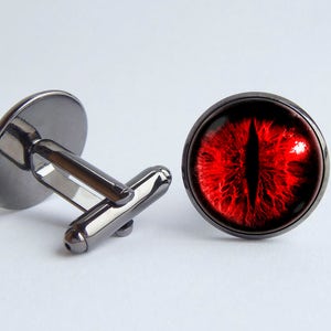 May include: A pair of cufflinks with a red eye design. The cufflinks are made of metal and have a black finish.