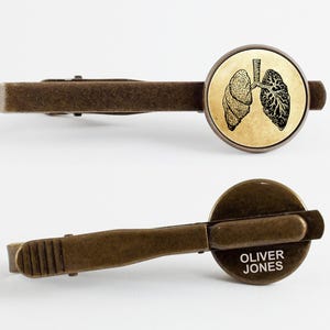 May include: A bronze tie clip with a round, antique-style image of a pair of lungs. The back of the clip is engraved with the name "Oliver Jones".