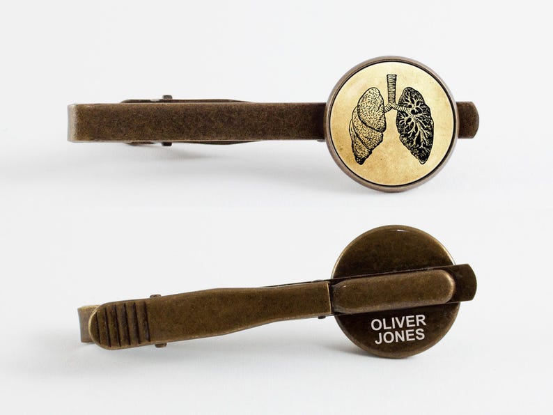 May include: A bronze tie clip with a round, antique-style image of a pair of lungs. The back of the clip is engraved with the name "Oliver Jones".