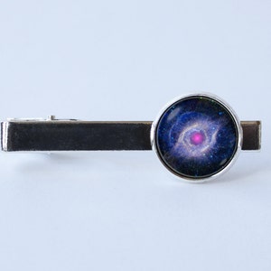 May include: A silver tie clip with a round glass cabochon featuring a colorful image of the Crab Nebula. The nebula is a deep space object with a pink center and blue and purple hues.