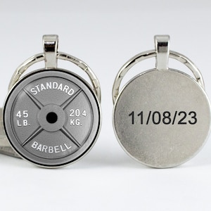 May include: Silver keychain with a round, gray weight plate featuring the text "STANDARD BARBELL 45 LB. 20.4 KG." on one side and the date "11/08/23" on the other side.
