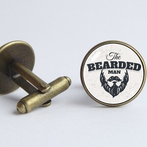 May include: A pair of cufflinks with a round, white background and black text that reads "The Bearded Man" with a black beard and mustache graphic. The cufflinks are made of brass and have a shiny finish.