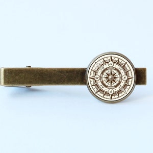 Peut inclure: Un fermo &agrave; cravate en laiton avec un motif de boussole rond. La boussole a un fond blanc avec des lignes et des marques brunes. Les points de la boussole sont &eacute;tiquet&eacute;s avec les lettres N, S, E et W.