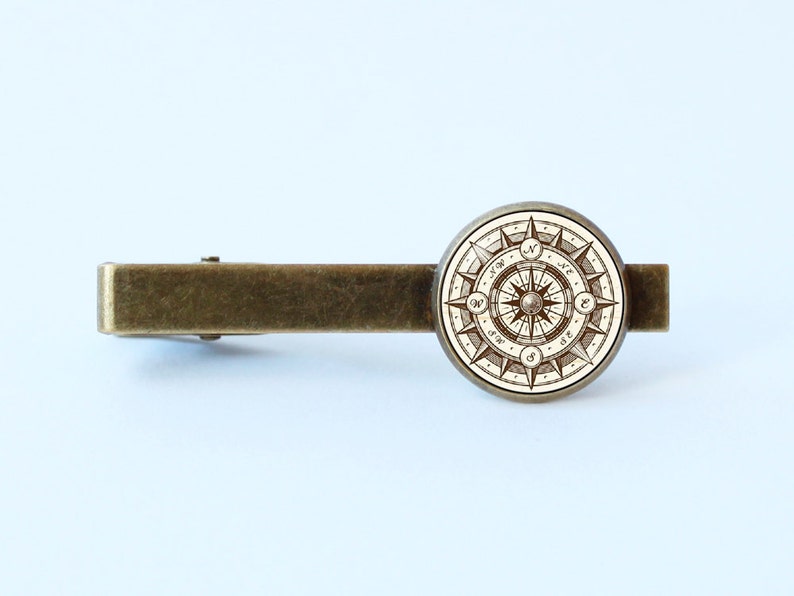 Peut inclure: Un fermo &agrave; cravate en laiton avec un motif de boussole rond. La boussole a un fond blanc avec des lignes et des marques brunes. Les points de la boussole sont &eacute;tiquet&eacute;s avec les lettres N, S, E et W.